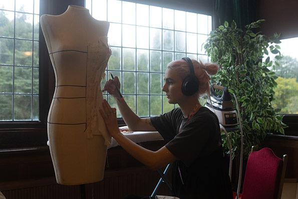 A person presenting as female, wearing headphones carefully pins and draws pattern lines onto a dress form in a studio space with large windows and greenery outside.
