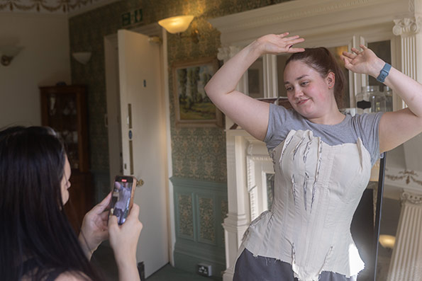 Two people presenting as female, one taking a picture of the other on her mobile phone. The scene and the picture on the mobile phone shows a carefully pinned and adjusted corset pattern being worn on top of a grey t-shirt and joggers by the person modelling it, in front of a mirror in a sunlit room. This image shows the model from the front.