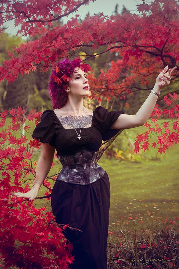 An elegant lady with dark pink hair is stood on a grass lawn next to a tree with red leaves. The figure is facing to the right. She is wearing a dark purple gothic dress and corset, looking into the distance.