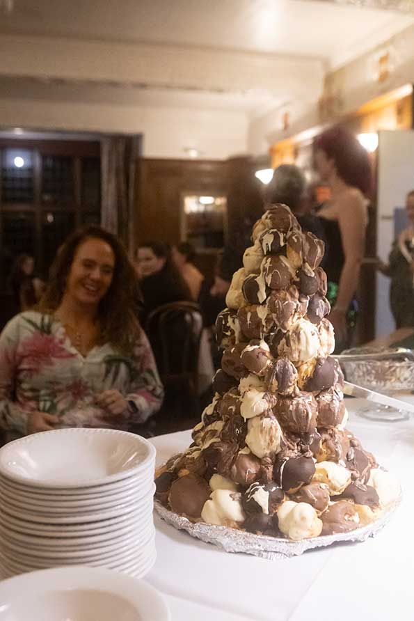 A photo showing a croquembouche, or croque-en-bouche, which is a French dessert consisting of choux pastry puffs piled into a cone and bound with threads of caramel.There is a table with a white linen tablecloth and white bowls. There are people who present as female seated in the background, enjoying dinner. The scene is not posed, indicating that it is a candid or impromptu shot.