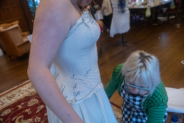 Two people presenting as female, one adjusting the corset pattern that the other person is modelling. They are in a brightly lit room with elegant furniture and a red patterned rug on a dark brown wooden floor. This image shows the model from the side. The scene is not posed, indicating that it is a candid or impromptu shot.