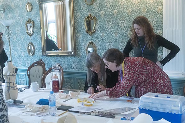 Three people presenting as female, taking part in a workshop to design and make corsets. The room is brightly lit, with blue and white floral patterned wallpaper, and silver mirrors. There is a table with a white linen tablecloth, an artist or designer's toolbox, and a small maquette on it. The scene is not posed, indicating that it is a candid or impromptu shot.
