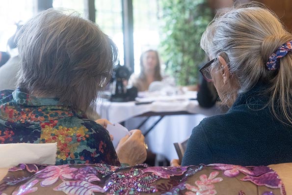 Two older people presenting as female. One appears to be reading, whilst the other is looking at some cards. They are in a brightly lit room with elegant furniture. This image shows the two people photographed from behind but the scene is not posed, indicating that it is a candid or impromptu shot.