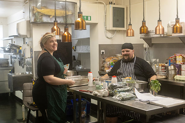 Two people, one presenting as female and one presenting as male. They are chefs in a kitchen, preparing a meal. This image shows the two people photographed smiling, but the scene is not posed, indicating that it is a candid or impromptu shot.