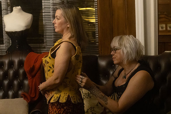 Two people presenting as female, one adjusting the corset that the other person is modelling. They are in a brightly lit room with elegant dark brown leather furniture in the style of a Chesterfield sofa. This image shows the model and corset designer from the side.