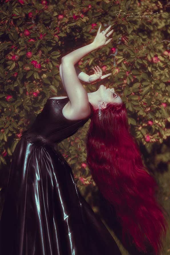 A posed photograph of a voluptuous, curvy model with a wrist tattoo, who presents as female. The model has long hair that is dyed a deep red colour, and is wearing a corset dress.There is an apple tree in the background. The model is leaning backwards so that her long red hair falls down to the ground.