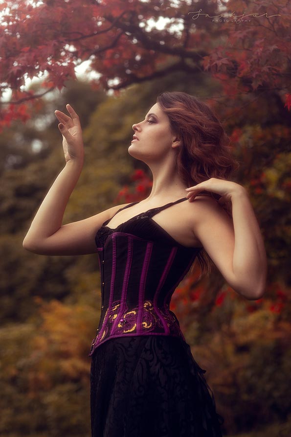 A posed professional photograph of a model in a black and purple velvet corset dress, who presents as female. The model is stood outside, and has bent arms.There is a tree with red leaves in the background, indicating that the photo was taken in the autumn.