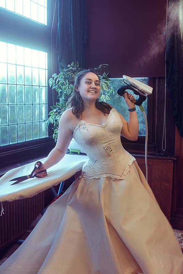 A smiling model wearing a white corset blank, and holding an iron and a pair of scissors or dress cutting shears. The model is stood by an ironing board, in front of a window. There is winter greenery outside the window.