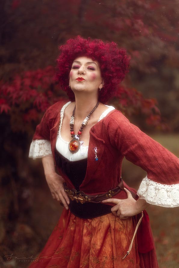 A posed professional photographer shot of a red haired model, who appears to present as a female, wearing a Restoration style corset dress. The model has red lips and rouge cheek makeup, and is stood outside, next to a tree. The tree has red leaves, indicating that the photo was taken in the autumn.