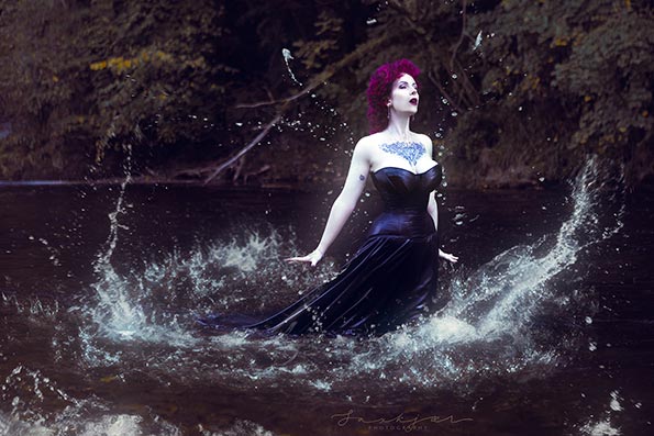 A posed professional photographer shot of a purple haired voluptuous model with tattoos, who appears to present as a female, wearing a black corset dress. The model is stood in a shallow river.