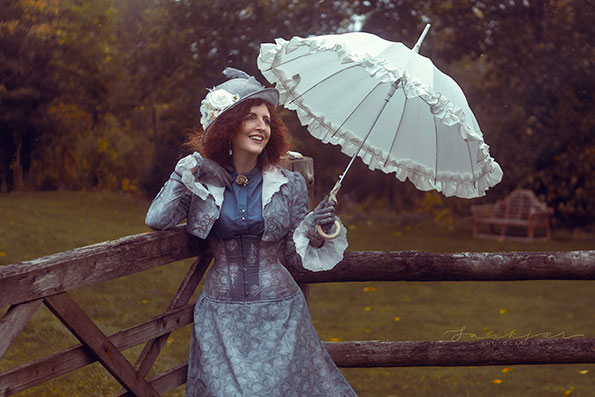 A professional photographer shot of a red haired model, who appears to present as a female, wearing a silver patterned Victorian style or steam punk corset dress. The model is outside, next to a wooden gate, and carrying an open parasol.