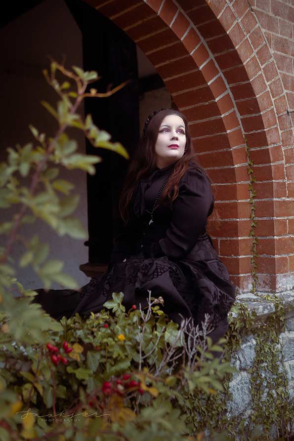 A professional photographer shot of a dark haired curvy and voluptuous model, who appears to present as a female, wearing a gothic style or steam punk black corset dress. The model is seated outside, next to a red brick archway.