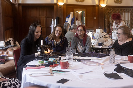 A group of people seated around a large table in a workshop setting, sewing and crafting together with fabric pieces, tools, mugs and sewing machines spread across the table. The scene is not posed, indicating that it is a candid or impromptu shot.