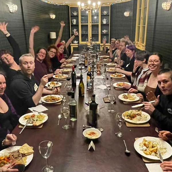 Meal time at The Corsetry Retreat March 2023. 14 people sat around a large table, 7 on each side. All eating an evening meal with glasses in front of them and bottles of wine on the table.