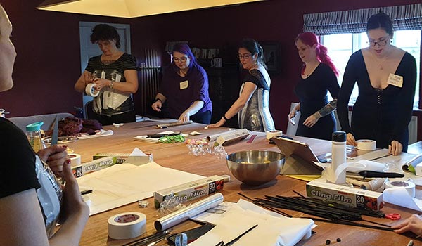 People at the March 2023 Corsetry Retreat. They are gathered around a large table and are building the base of a corset pattern with tape and cling-film.