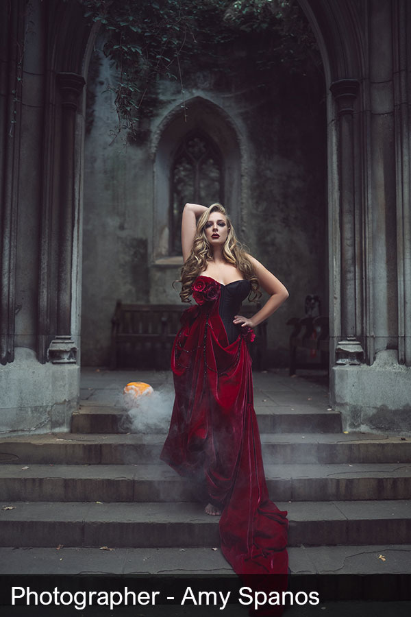 A person wearing a black corset with a blood red drape and matching skirt. She has long, wavey blonde hair and is stood in front of a large church doorway.