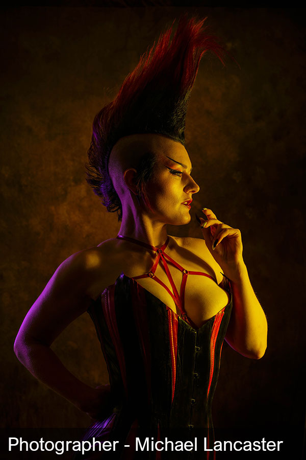 A dark black and red lit image of a person wearing a red stripy corset and facing to the right. They have a huge Mohican hair style. The photographer is Michael Lancaster.
