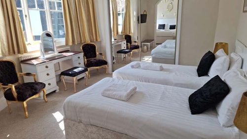 A photograph of a bedroom with 2 single beds, on the far wall there is a mirrored wardrobe and the mirror shows that out of shot there is an additional double bed. Next to the mirror there is a white dressing table with black and gold chairs.