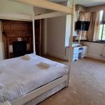 A photograph of a bedroom with a 4-poster bed with white sheets and a dark wood fireplace behind it. There is also a white dressing table on the far wall.