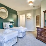 A photograph of a twin bedroom with dark green walls with a round mirror above the beds, opposite the beds there is a dark wood dressing table and mirror.