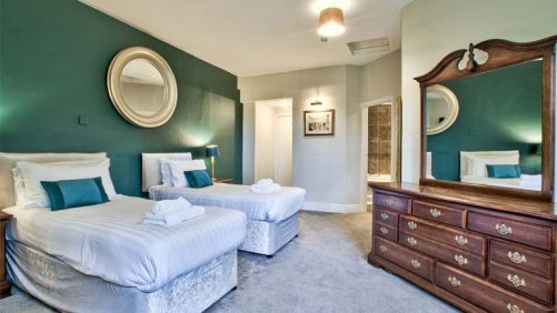 A photograph of a twin bedroom with dark green walls with a round mirror above the beds, opposite the beds there is a dark wood dressing table and mirror.