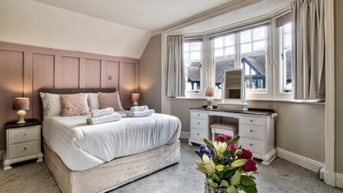 A photograph of bedroom with pink wood panelling behind a bed with white bedsheets. To the right of the bed there is bay window looking out onto a courtyard, in front of the window is a white dressing table with a mirror on top and in the foreground of the picture are some white and red flowers.