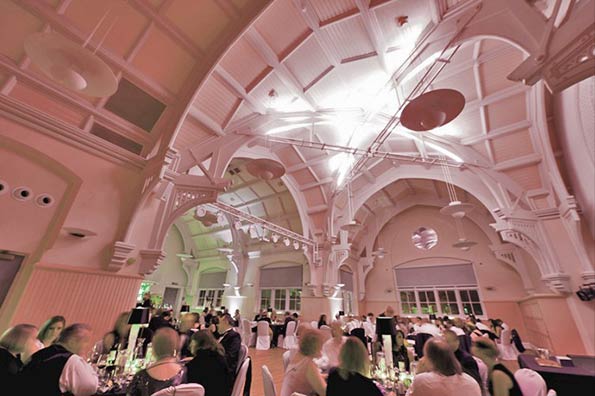 Inside a large conference room where it is lit in pink and everyone is sat around tables in suits and dresses.