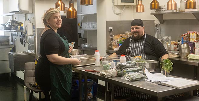 Two people, one presenting as female and one presenting as male. They are chefs in a kitchen, preparing a meal. This image shows the two people photographed smiling, but the scene is not posed, indicating that it is a candid or impromptu shot.