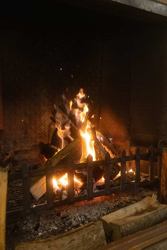 A fireplace, with a lit, roaring fire. There are logs in front of the metal fire grate, and burning logs inside the fireplace itself.