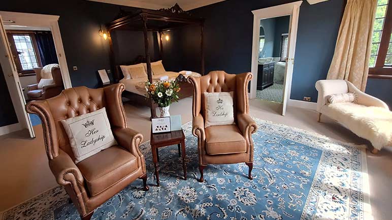A luxury bedroom with en suite bathroom, four poster bed, leather wing-back chairs and elegant furnishings.